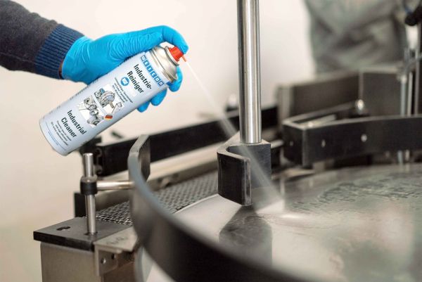 A person wearing a blue glove sprays WEICON Industrial Cleaner onto a rotating metal turntable on a machine.
