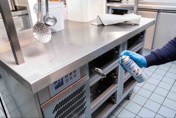 A person wearing a blue glove sprays WEICON Bio-Lube Fluid onto the internal track mechanism of a commercial stainless steel kitchen counter unit.