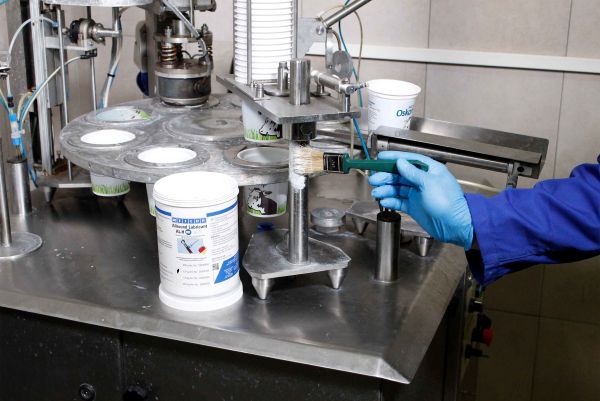 A person wearing a blue glove uses a paintbrush to apply lubricant from a WEICON container onto a vertical metal component of a packaging machine.