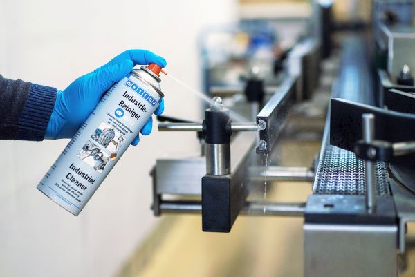 A person wearing a blue glove sprays WEICON Industrial Cleaner onto a metallic conveyor belt mechanism.