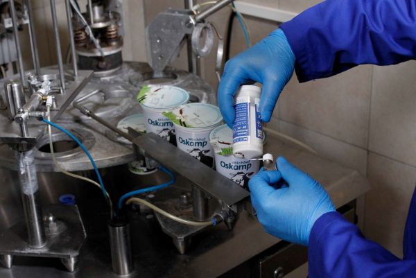A person wearing blue gloves applies lubricant from a small WEICON bottle onto a machine component near a dairy packaging station.