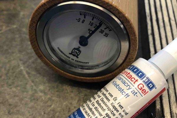 A tube of WEICON Contact Gel cyanoacrylate adhesive lies next to a round analog gauge framed in wood.