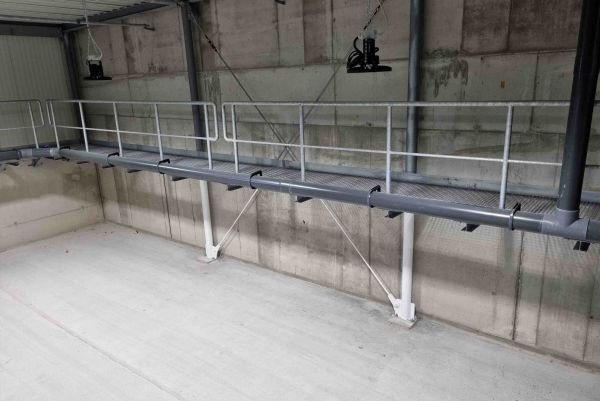 An elevated metal catwalk with safety handrails and a dark pipe runs along a concrete wall inside an industrial facility.