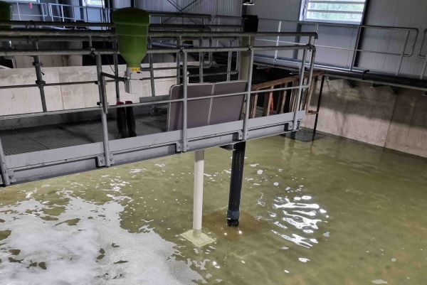 A elevated metal walkway with safety railings extends over a large water treatment tank with foamy water.